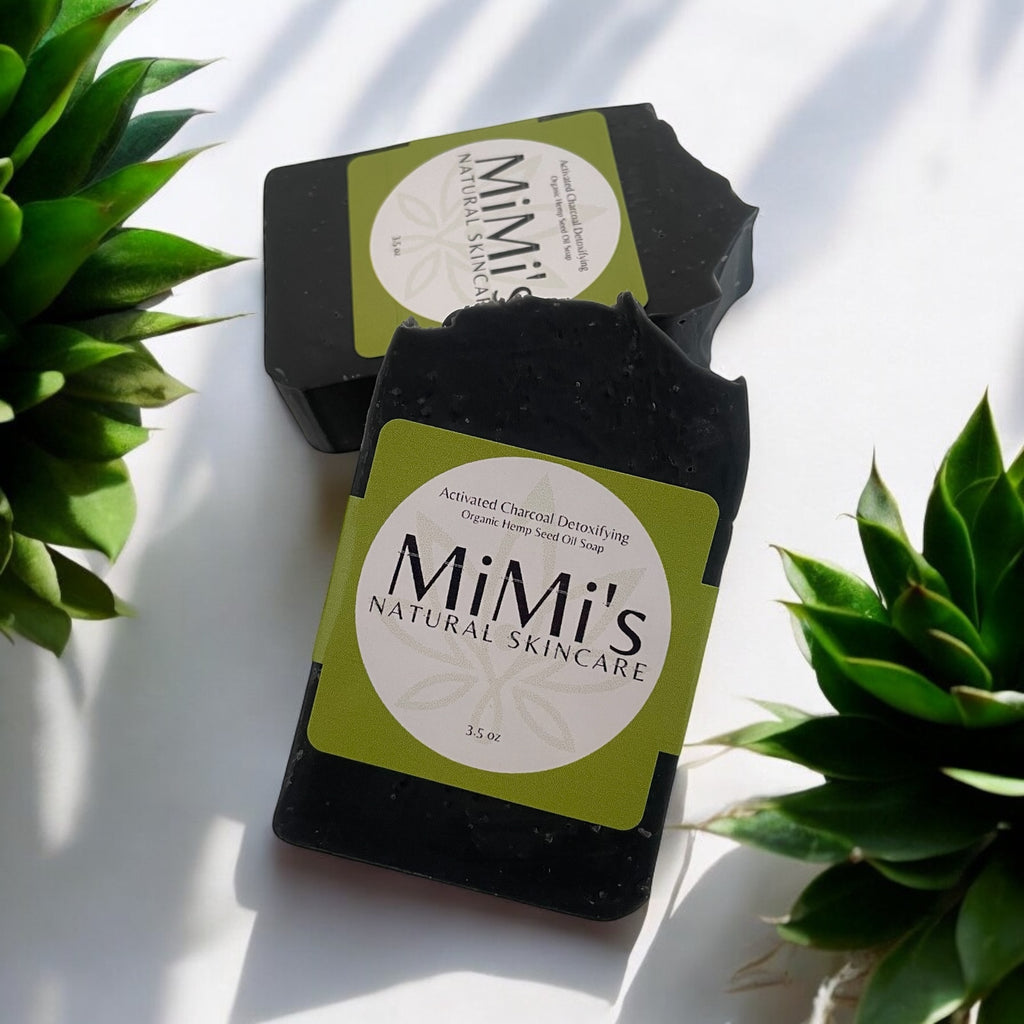 Two bars of activated charcoal detoxifying face soap with packaging that reads 'MiMi's Natural Skincare'. The soap bars are black and placed against a bright background with green foliage.